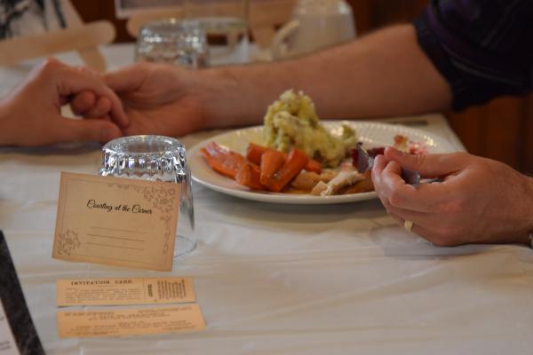 A couple holds hands while eating a roast dinner.  Next to the plate is an invitation card for the event.  