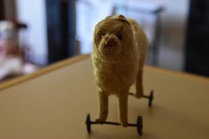 A small toy dog with metal wheels on its legs faces the viewer. The toy's fur is worn short, and its nose is ripped. The toy looks very old.