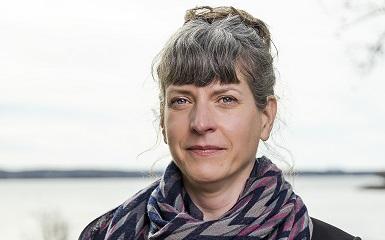 A head-and-shoulders image of a woman with dark brown hair with grey streaks. Her hair is tied back behind her head and she is wearing a large scarf wrapped around her neck. She has a slight smile and is looking at the viewer. In the background is a green lawn and beyond that is a body of water.