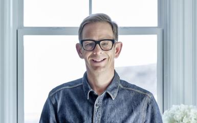 A man with glasses wearing denim clothes, standing by the window