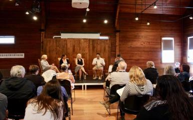 An event hosted at Beaconsfield Carriage House. There are 3 speakers on the main stage, as well as attendees sitting on rows looking at the stage.