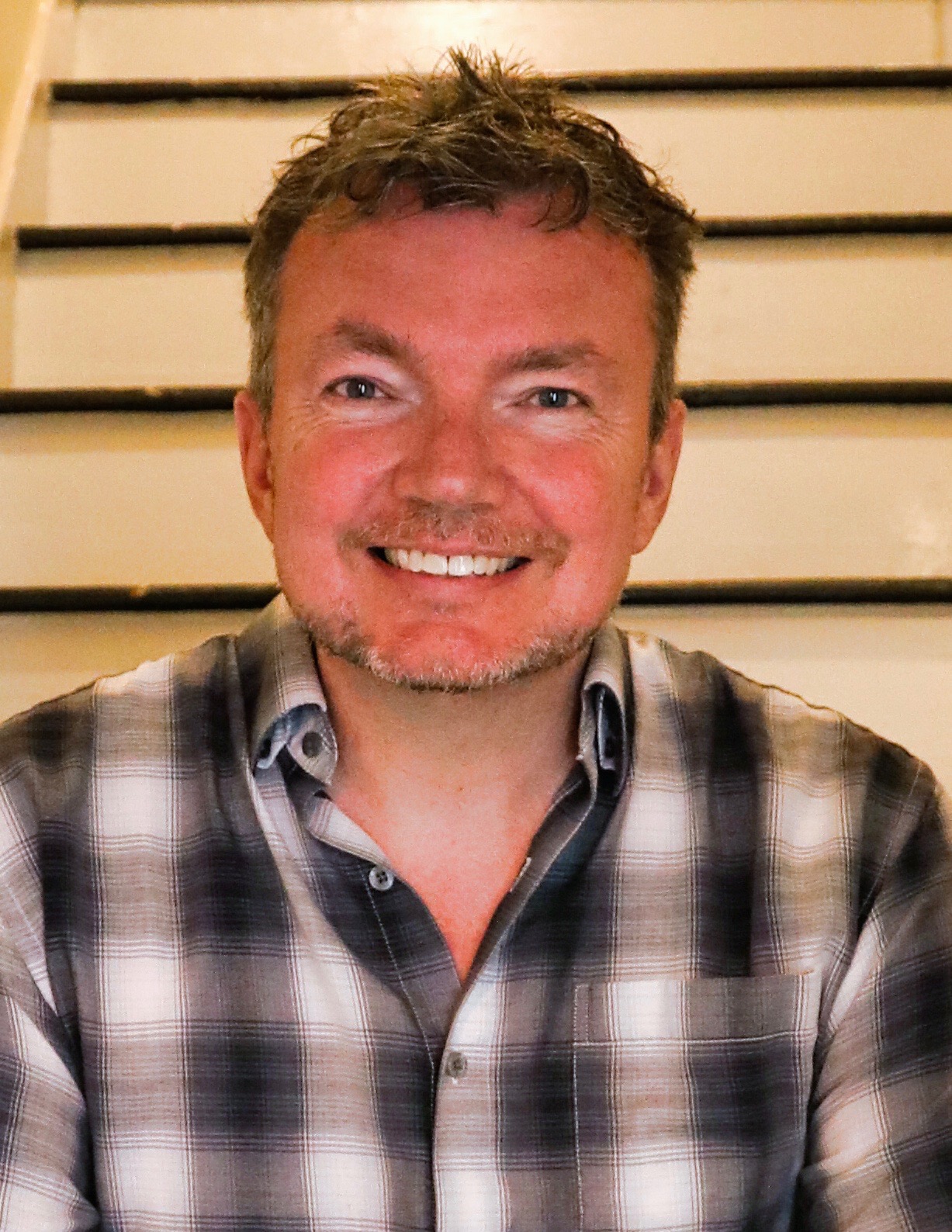 A headshot of a smiling man wearing a plaid shirt.