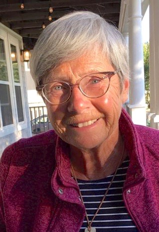 A smiling woman with glasses sits in a bright windowed room. 