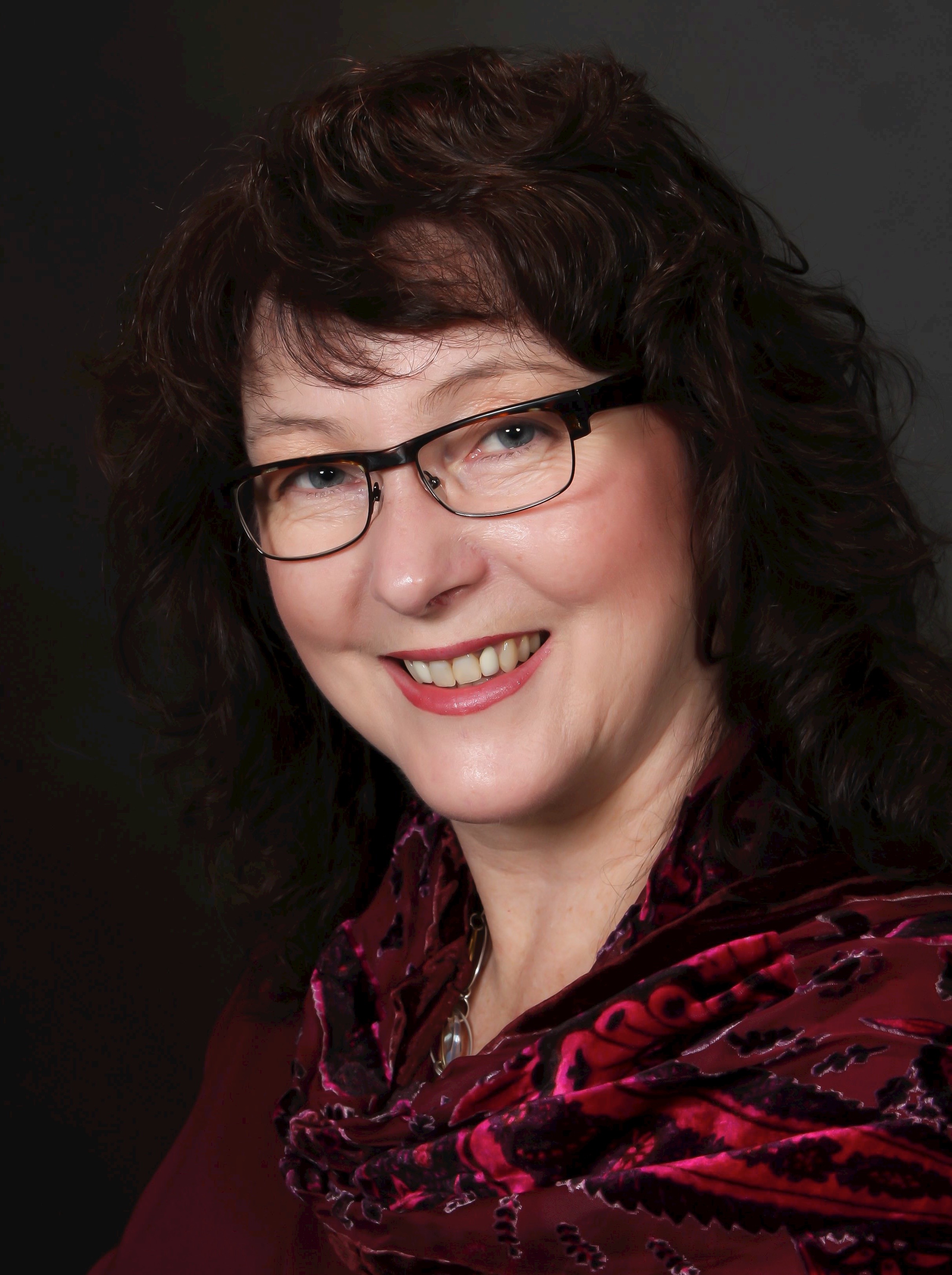 A headshot of a woman with dark hair sitting infront of a dark grey background.