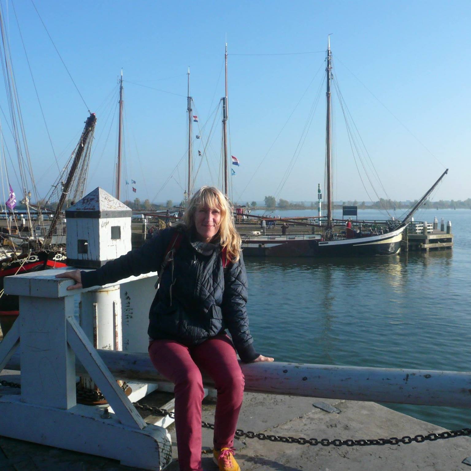 A woman sits on the railing of a marina with ships in the background.