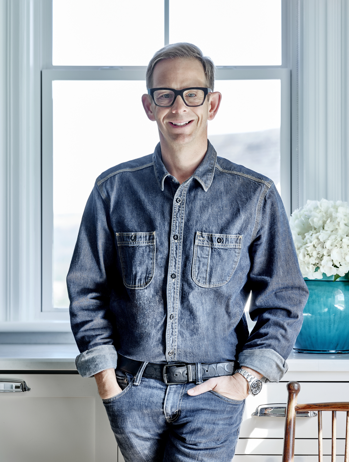 A man with glasses and denim clothes standing by a window 
