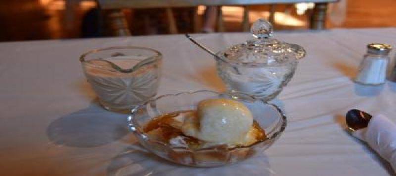 A bowl of bread pudding with ample caramel sauce and ice cream sits on a set table with a bowl of sugar and cream behind it. 