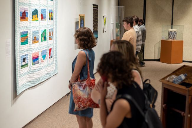 A group of sex people stand observing items on display in a gallery. Two women in the foreground observe something out of shock. A woman in the midground looks at a quilt hanging on a wall. Next to her a man looks at a stained glass artwork. On the background two men look at an item that is out of view. 