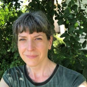 A woman wearing a green shirt stands in front of green leafy trees