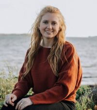 A woman with long curly hair sits on a rock in front of an unknown body of water