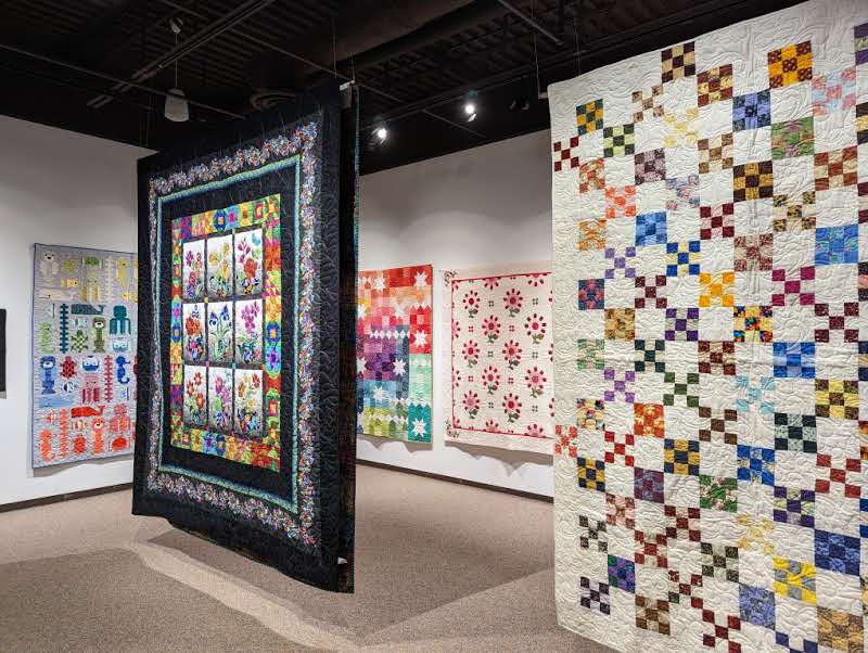 Quilts of Covid Exhibit at Eptek Art and Culture Centre. On the photo are 5 colorful quilts hung on the wall and from the ceiling of Eptek.