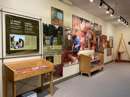 Exhibition panels including text and photos of rocks, archaeologists digging, site plots. Two wooden cases displaying indiscernible artifacts.