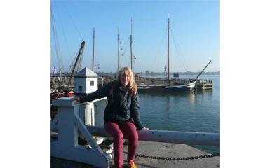 A woman with blonde hair sits on the railing of a marina with boats in the background