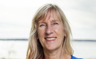 A head-and-shoulders image of a woman with long blonde hair who is wearing a blue shirt. She is looking at the viewer and smiling. In the background is a green lawn and beyond that is a body of water.