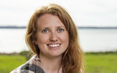 A head-and-shoulders image of a woman with long blonde hair that goes past her shoulders. She is wearing a black and white plaid scarf around her shoulders. She is looking at the viewer and smiling. In the background is a green lawn and beyond that is a body of water.