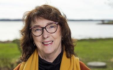 A head-and-shoulders image of a woman with shoulder length brown hair who is wearing glasses and a yellow scarf that hangs around her neck. She is looking at the viewer and smiling. In the background is a green lawn and beyond that is a body of water.